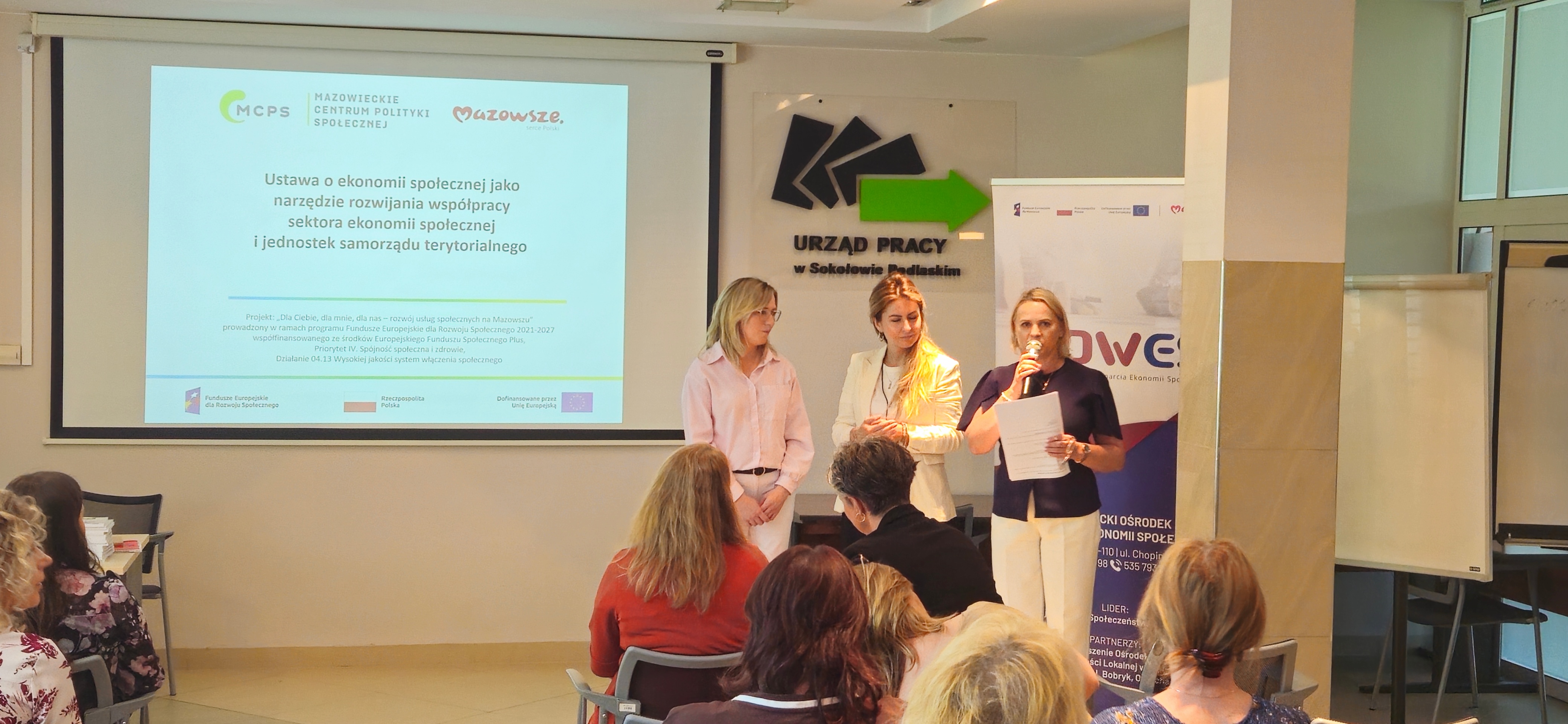 Trzy kobiety prowadzące prezentację podczas spotkania w sali konferencyjnej; jedna z nich przemawia do mikrofonu, trzymając dokument. W tle widoczny ekran z prezentacją dotyczącą ustawy o ekonomii społecznej oraz logo instytucji, a przed nimi siedzą uczestnicy wydarzenia.