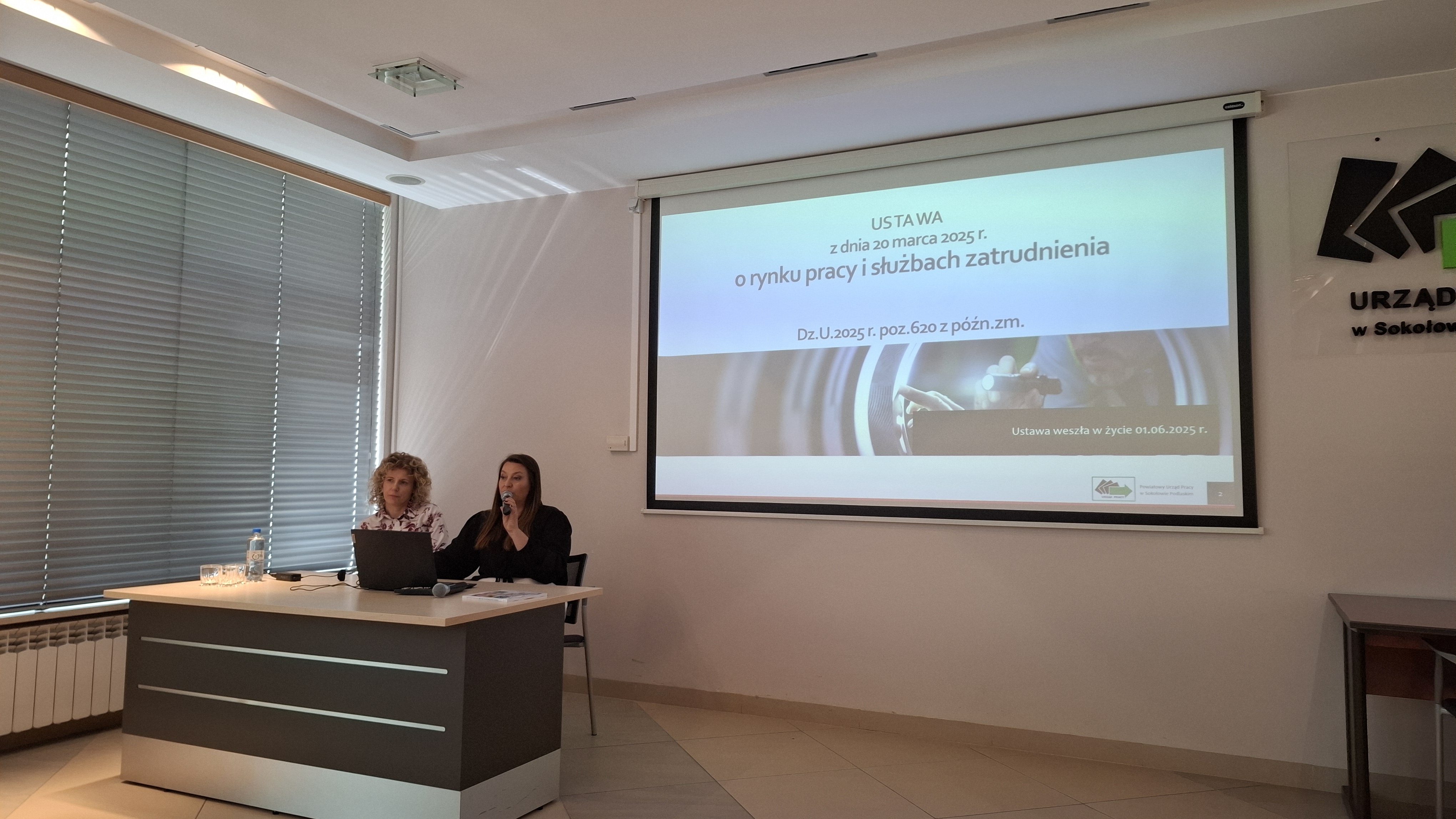 Dwie prelegentki siedzące przy stole w sali konferencyjnej podczas prezentacji; jedna z nich przemawia do mikrofonu, a na stole znajduje się laptop. W tle widoczny ekran z wyświetloną prezentacją dotyczącą ustawy o rynku pracy i służbach zatrudnienia.